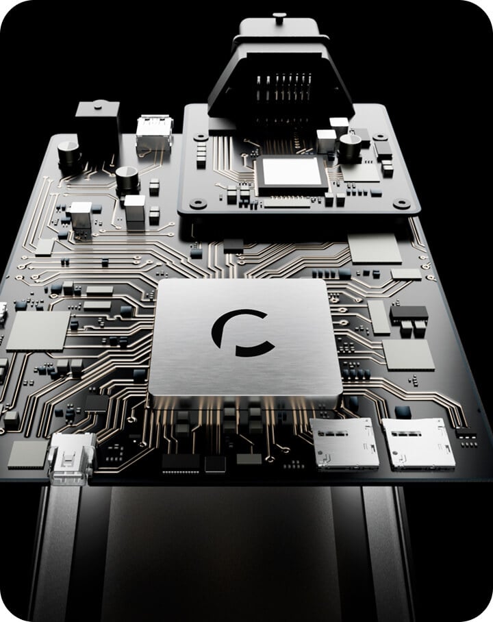 Close-up view of a computer circuit board with various electronic components, ports, and a metallic central chip displaying a stylized "C" logo, set against a dark background.